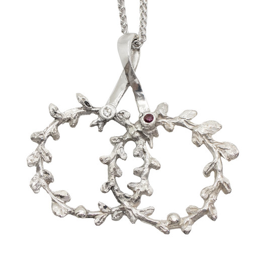 Silver necklace with two intertwined olive leaf crowns, adorned with a diamond and a ruby.