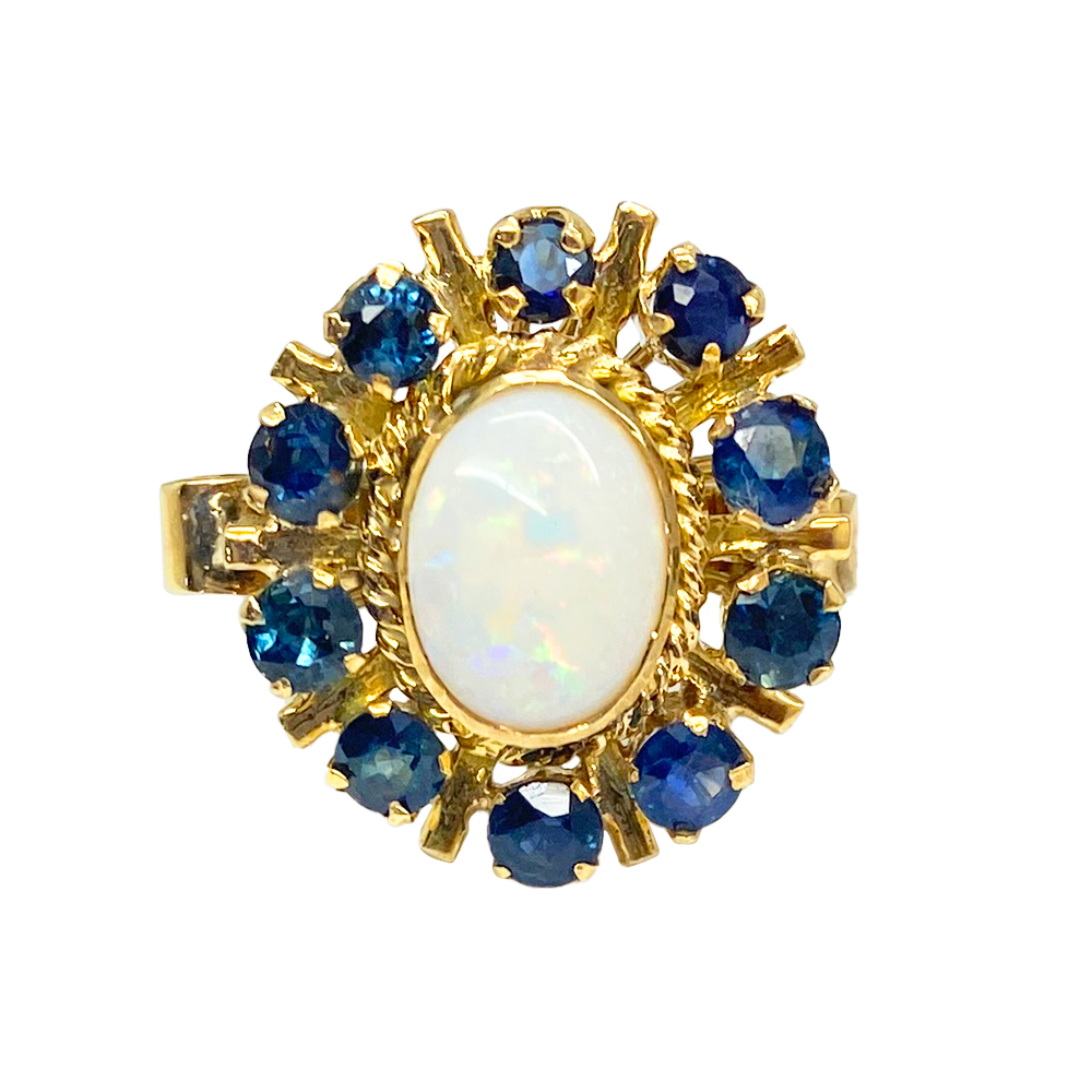 Gold ring featuring a large oval opal surrounded by a cluster of blue sapphires. The ring has a delicate, open-work design.