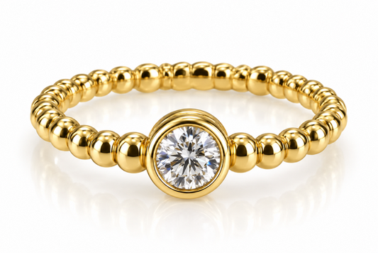 A gold beaded stretch ring featuring a large round bezel-set clear gemstone in the center, displayed on a clean white reflective surface.