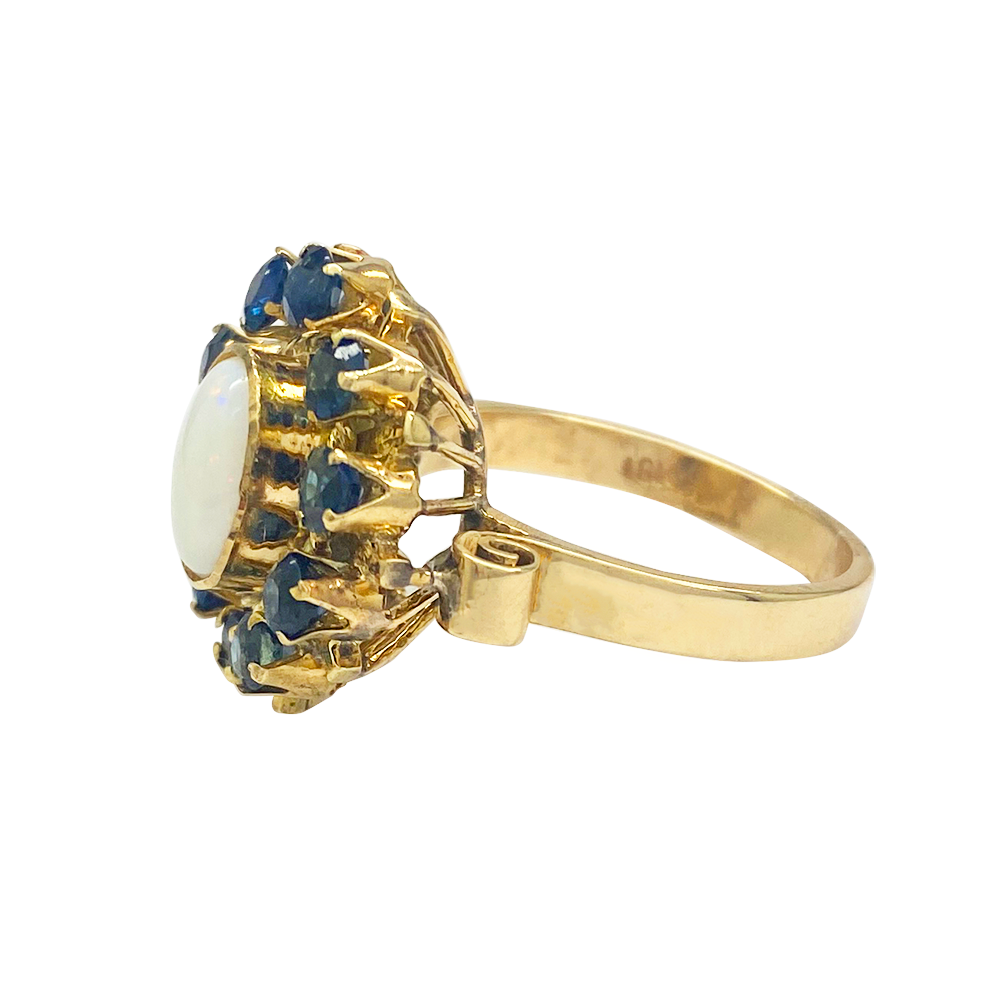Gold ring featuring a large oval opal surrounded by a cluster of blue sapphires. The ring has a delicate, open-work design.