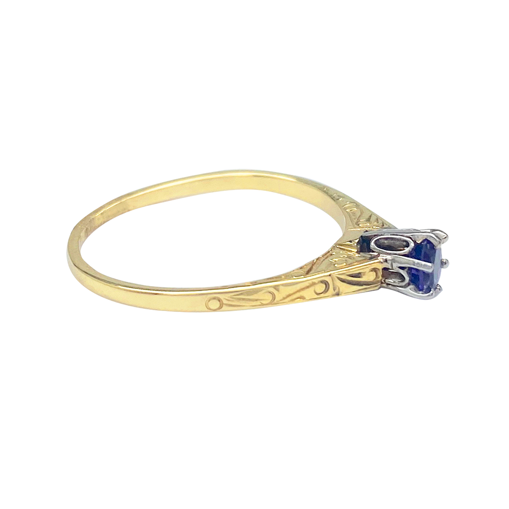 Gold ring with a round sapphire set in a four-prong setting. The band is engraved with intricate scrollwork.