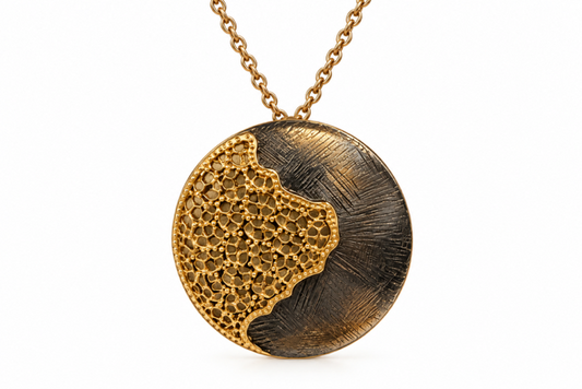 A round pendant necklace featuring a split design: one half is textured gold with a honeycomb pattern, and the other half has a brushed dark metallic finish. The pendant hangs from a gold link chain against a plain white background.
